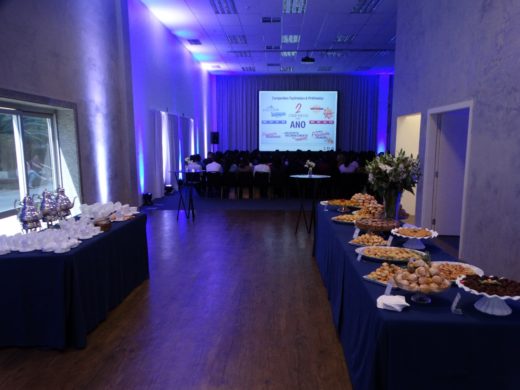 Salão de convenção corporativa com palestra ao fundo e mesas de buffet e coffee break preparadas para os convidados.
