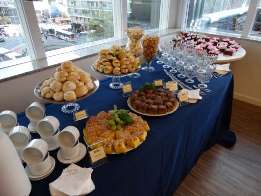 Mesa de coffee break para empresas com mini sanduíches, bolos e garrafas de café em sala de reunião.