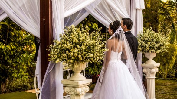 Noiva de costas com véu longo e noivo no altar durante cerimônia de casamento ao ar livre.